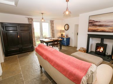 A living room with a beige sofa with a red throw a black wood stove with fire a wooden dining table with chairs and a dark wardrobe at Rhytty Bach in Ty Croes near Rhosneigr