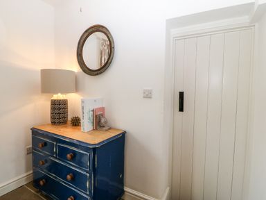 A corner with a blue wooden dresser a lamp books and decorative items on top and a round mirror on the wall at Rhytty Bach in Ty Croes near Rhosneigr