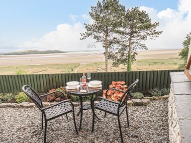 An outdoor patio area with a small round table and two chairs overlooking a sandy beach and grassy area at Oversands Cottage in Grange-Over-Sands