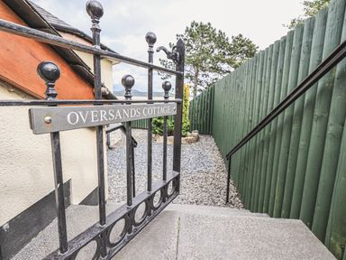 A metal gate with Oversands Cottage sign next to stone steps and gravel path at Oversands Cottage in Grange-Over-Sands