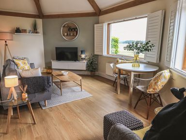 A living room with sofa, coffee table, TV, round dining table with chairs, and large window at Oversands Cottage in Grange-Over-Sands