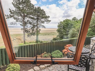 A view through an open wooden window of a garden with chairs and table and a fence with trees and a beach beyond at Oversands Cottage in Grange-Over-Sands
