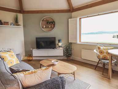 A living room with a gray sofa and yellow pillows a wooden coffee table TV on a wall unit and a window with a view at Oversands Cottage in Grange-Over-Sands