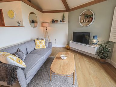 A living room with a gray sofa yellow pillows wooden coffee table tv on a white floating cabinet and wooden floor at Oversands Cottage in Grange-Over-Sands
