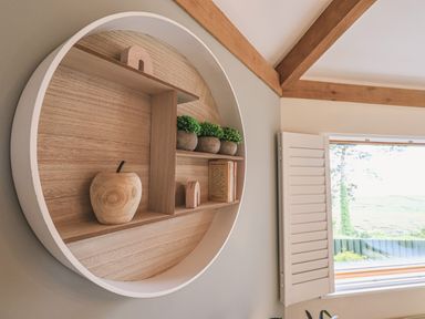 A round wooden wall shelf with small decorative items and plants near a window with white shutters at Oversands Cottage in Grange-Over-Sands