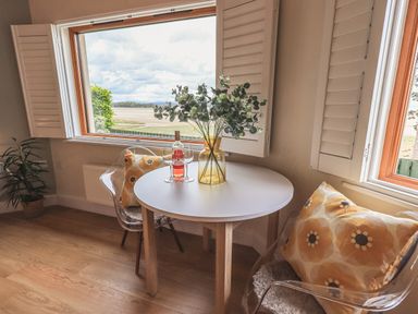 A small round table with a vase of greenery and a bottle with two glasses by two transparent chairs with patterned cushions near large windows at Oversands Cottage in Grange-Over-Sands