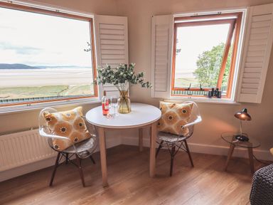 A small dining area with two chairs around a round table and a scenic view through two corner windows at Oversands Cottage in Grange-Over-Sands