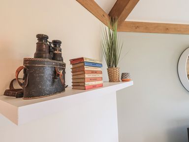 A white wall shelf with vintage binoculars in a leather case stacked books a potted plant and a small decorative pot at Oversands Cottage in Grange-Over-Sands