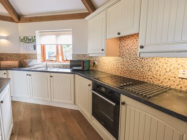 A kitchen with white cabinets a black countertop a built in oven an electric stovetop a window and a dish rack at Oversands Cottage in Grange-Over-Sands