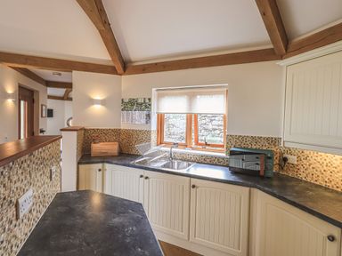 A kitchen with white cabinets black countertops a window and wooden ceiling beams at Oversands Cottage in Grange-Over-Sands