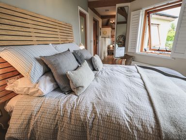 A bedroom with a bed with patterned bedding and multiple pillows next to an open window at Oversands Cottage in Grange-Over-Sands