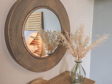 A round mirror on a wall reflecting a bed and a glass vase with dried flowers on a wooden surface at Oversands Cottage in Grange-Over-Sands