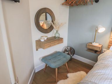 A corner of a bedroom with a blue stool a round mirror a small wall shelf with bowls a vase with dried flowers and a bedside table with a lamp at Oversands Cottage in Grange-Over-Sands