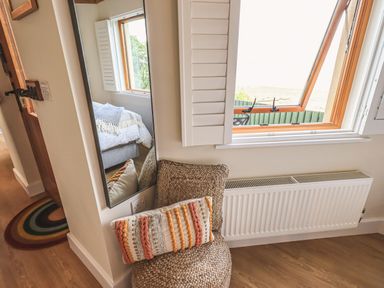 A corner of a room with a window radiator a wicker chair with a decorative pillow and a tall mirror reflecting a bed at Oversands Cottage in Grange-Over-Sands