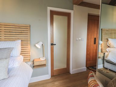 A bedroom with a bed and pillows next to a wooden door and a wall-mounted nightstand with a lamp at Oversands Cottage in Grange-Over-Sands