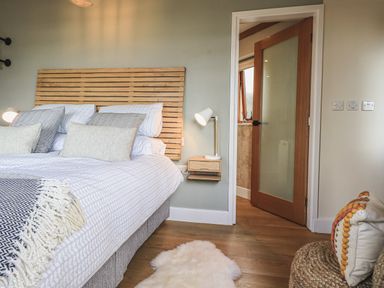 A bedroom with a wooden headboard bed with pillows a bedside table with lamp an open door showing a bathroom a woven chair with a colorful cushion and a rug on wooden floor at Oversands Cottage in Grange-Over-Sands