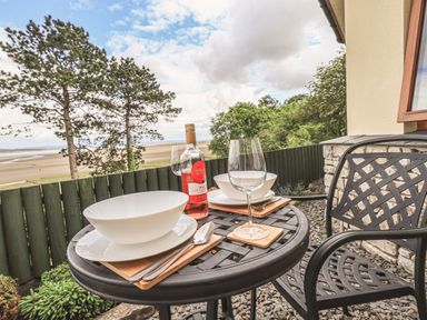 An outdoor patio table with two place settings wine glasses and a bottle with trees and beach in the background at Oversands Cottage in Grange-Over-Sands