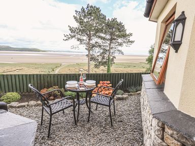 A small outdoor dining area with two metal chairs and a round table set with bowls and wine overlooking a beach landscape at Oversands Cottage in Grange-Over-Sands