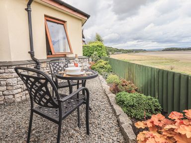 An outdoor patio with a small black metal table set for two and a view of a green fence and open landscape at Oversands Cottage in Grange-Over-Sands