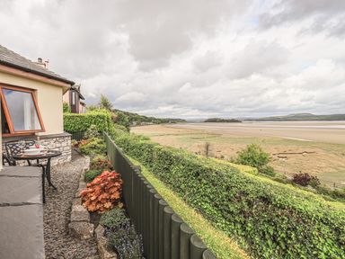 A small outdoor patio with a table and chairs next to a house overlooking a grassy beach and water at Oversands Cottage in Grange-Over-Sands
