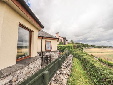 An outdoor patio area with a table and chairs next to a house overlooking a grassy area and a beach at Oversands Cottage in Grange-Over-Sands