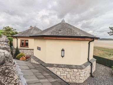 Exterior view of a round cottage with stone and cream walls and a shingled roof near a beach at Oversands Cottage in Grange-Over-Sands