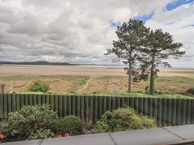 A view of a coastal landscape with sand, greenery, trees, and a cloudy sky at Oversands Cottage in Grange-Over-Sands