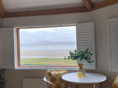 A room with a round white table and yellow flowered cushions near a window with shutters at Oversands Cottage in Grange-Over-Sands
