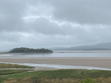 A view of a coastal landscape with grassy marshland in the foreground sandy beach and water leading to an island and hills under a cloudy sky at Oversands Cottage in Grange-Over-Sands