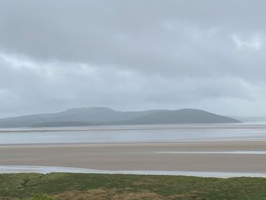 A coastal scene with green grass in the foreground a sandy shore and water with hills in the background under a cloudy sky at Oversands Cottage in Grange-Over-Sands
