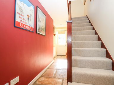A hallway with stone floor stairs with carpet and framed pictures on a red wall at 1 The Lane in Chipping Norton
