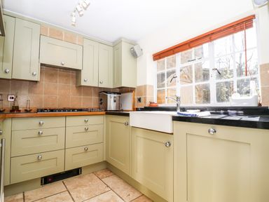 A kitchen with olive green cabinets, a black countertop, a white farmhouse sink, a gas stove, and a window with wooden blinds at 1 The Lane in Chipping Norton
