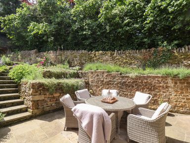 An outdoor patio with a round table and six cushioned wicker chairs next to stone steps and garden walls at 1 The Lane in Chipping Norton