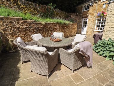 An outdoor patio with a round glass table and six wicker chairs with cushions at 1 The Lane in Chipping Norton
