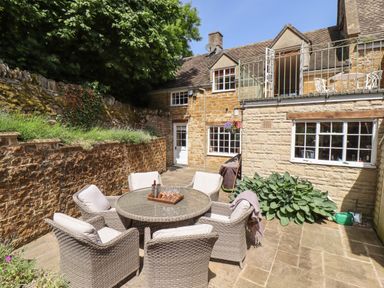 An outdoor patio with a round glass table, six cushioned wicker chairs, stone walls, plants, and a two-story stone house at 1 The Lane in Chipping Norton