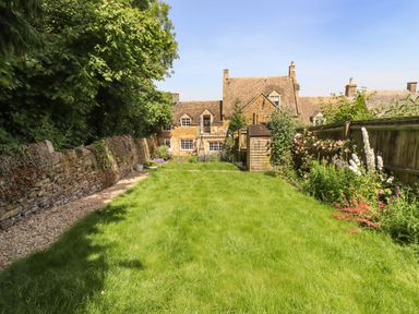 A garden with lawn bordered by flowers and shrubs with stone wall and wooden fence and stone house in the background at 1 The Lane in Chipping Norton