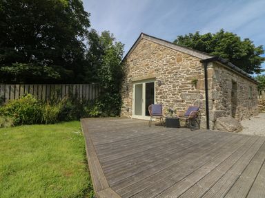 A stone building with a wooden deck and two chairs next to a grassy lawn at The Reading Room in Seworgan near Constantine