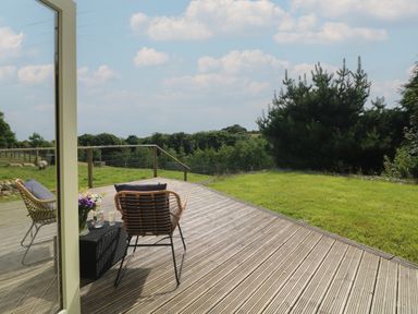 A wooden deck with two chairs and a small table with flowers and drinks overlooking grass and trees at The Reading Room in Seworgan near Constantine