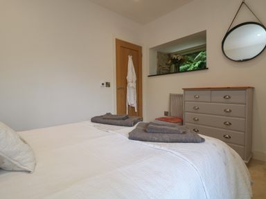 A bedroom with a bed covered in white bedding gray towels a grey dresser with drawers a chair with a red cushion and a round mirror on the wall at The Reading Room in Seworgan near Constantine