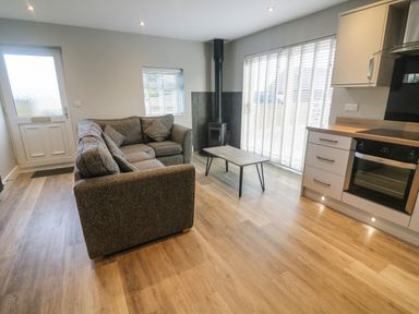 A living area with a grey corner sofa, coffee table, wood stove, and kitchen with wooden floor at White Stones in Goadsbarrow near Roose