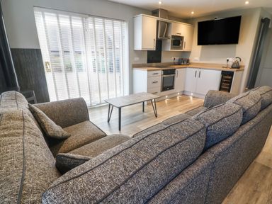 A kitchen and living area with a gray sofa a coffee table a wall-mounted TV and sliding glass doors at White Stones in Goadsbarrow near Roose