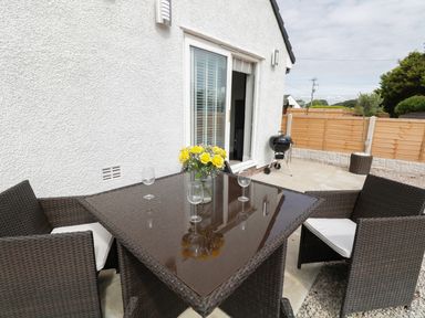 An outdoor patio with a woven table and chairs a vase with yellow flowers and glasses a barbecue near a sliding door at White Stones in Goadsbarrow near Roose