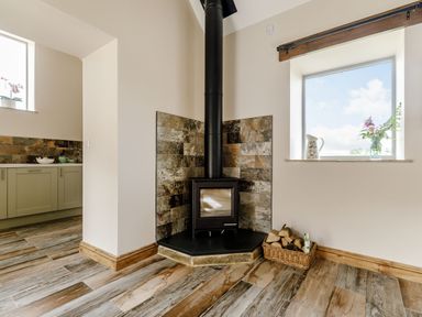 A corner fireplace with a wood stove and basket of firewood next to a window with flowers at Foxglove in Goathland