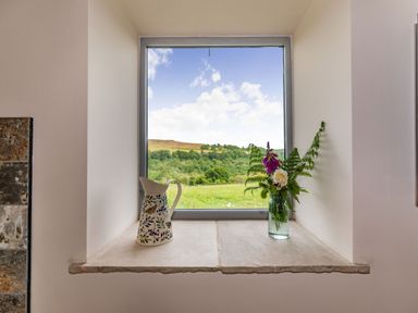 A window with a ceramic jug and a vase with flowers on the sill overlooking a green landscape at Foxglove in Goathland