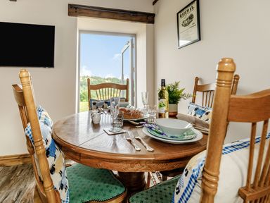 A dining area with a round wooden table set with plates glasses and cutlery and wooden chairs with cushions near an open door with a view outside at Foxglove in Goathland