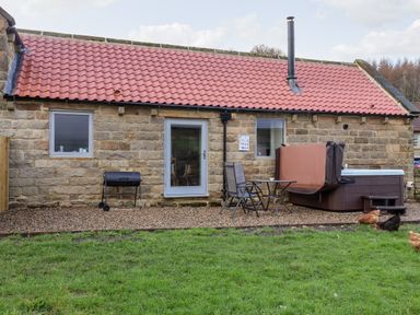A stone building with a red tiled roof a glass door two windows a black barbecue grill two chairs a small table a covered hot tub and chickens on a grassy area at Foxglove in Goathland