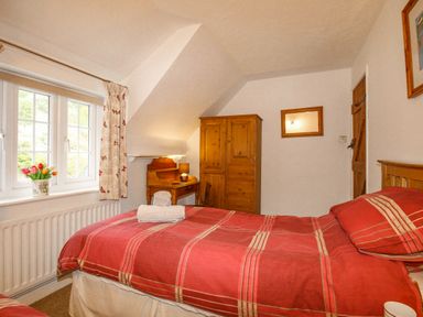 A bedroom with a red bedspread a wooden wardrobe a small desk and a window with floral curtains at Cajun Cottage in West Lulworth
