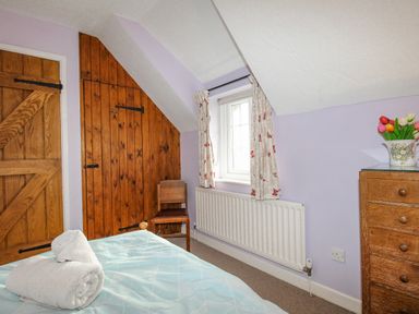 A bedroom with a wooden chair wooden doors a window with curtains a heater a chest of drawers and two towels on a bed at Cajun Cottage in West Lulworth