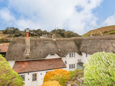 A thatched roof cottage with chimneys surrounded by bushes and hills at Cajun Cottage in West Lulworth
