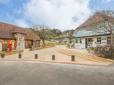 A paved outdoor area with a brick building and a blue and white striped building with picnic tables near trees at Cajun Cottage in West Lulworth